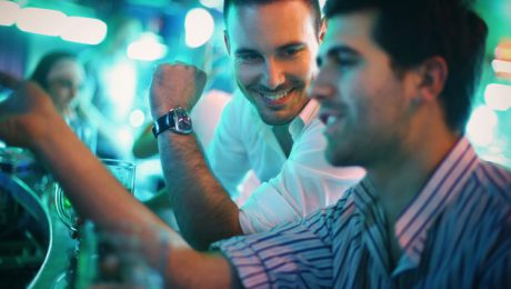 Two early 30's caucasian men having beer in a bar on weekend night. Having casual conversation and relaxing after working week.Sitting at bar counter and toasting with beer mugs. Added vignette/